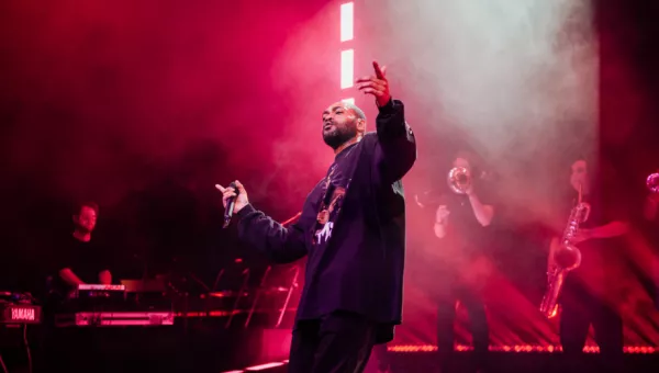 Kano Performing at the O2. Kano is standing on the stage, posing with his arms apart with red and purple lights behind him
