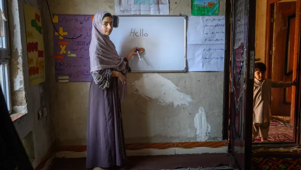 Young girl wearing a maxi dress and grey scarf stands in front of a whiteboard with the 'Hello' written.