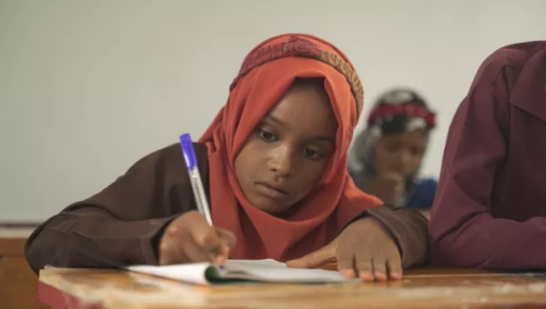 Young girl sat on a table with pen in her hand writing in her jotter.
