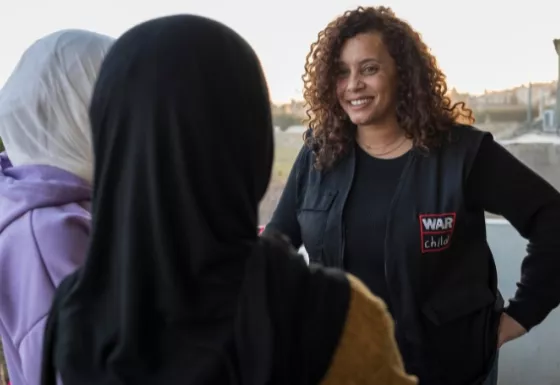 War Child worker wearing a branded vest smiling at two young girls wearing hijab.