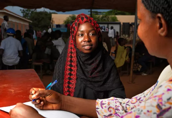 Young Sudanese girl, wearing a red and black coloured hijab, sat down on a bench talking to a War Child partner