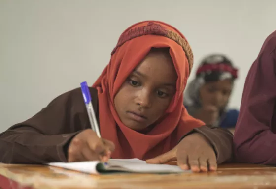 Young girl sat on a table with pen in her hand writing in her jotter.