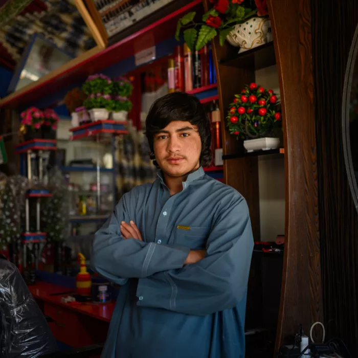 Young Afghan teen boy wearing traditional blue Afghani, hands crossed standing inside a shop.