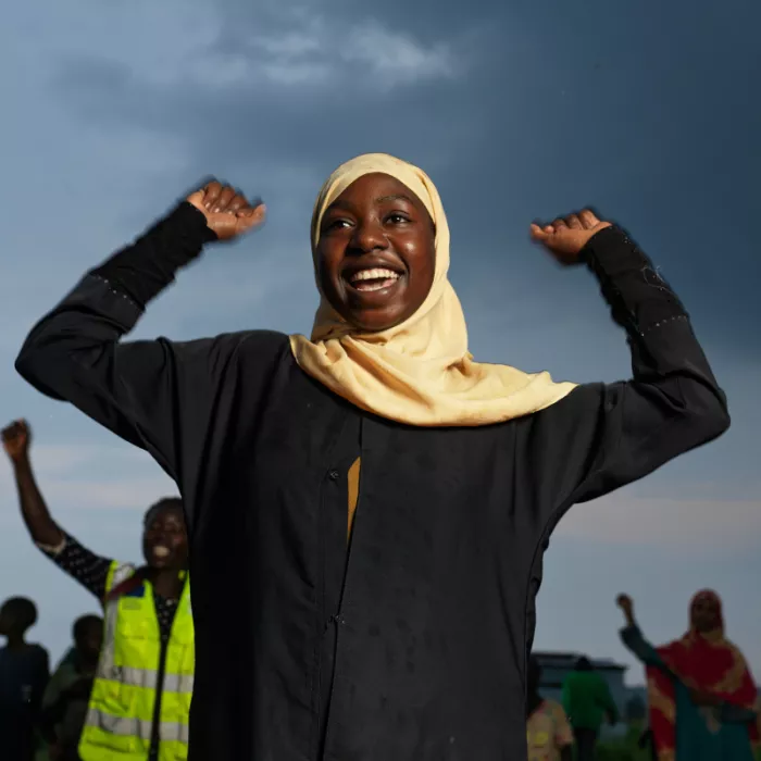 Young Sudanese girl wearing a yellow hijab, smiling with her hands in the air.